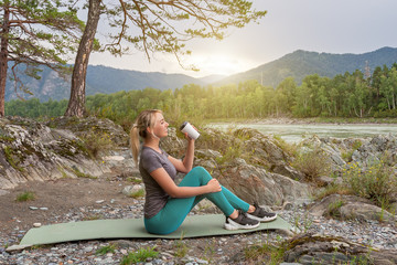 Blond beautiful girl sitting on the mat on the stone ground drinking coffee or tea, resting after sport outdoors in the mountains near the river. Workout and picnic in nature.