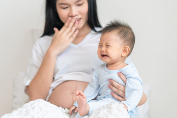 Asian male toddler sitting with his pregnant mother in bedroom. Crying face.