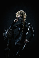 Fototapeta na wymiar Young cyborg woman in leather suit and a gas mask leaning on a headlight