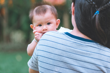 Close up of young asian mother sitting on bench with her male toddler with green trees and garden in background. Toddler looking at camera. Sucking finger with curious look.