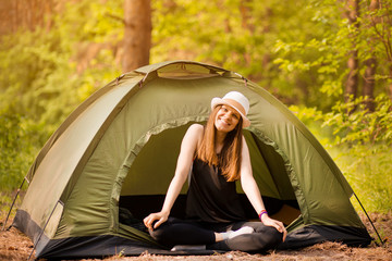 Happy woman in hat sits in tent around forest under sunset light sky enjoying leisure and freedom.