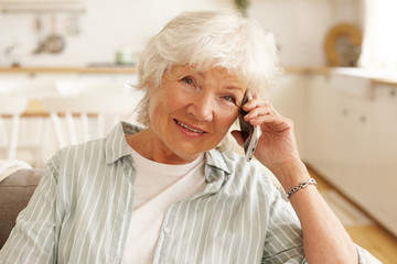 Elderly mature European woman in striped shirt having phone conversation via online application using free wireless high speed internet connection at home, looking at camera with cheerful smile
