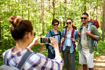 travel, tourism and hike concept - group of friends with backpacks being photographed by woman with smartphone in forest