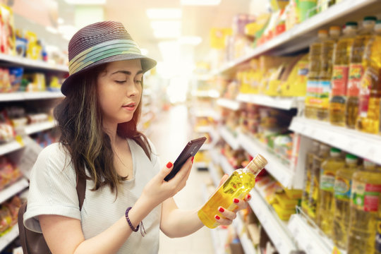 A young beautiful woman holds a bottle of oil in her hand and takes a picture of her on a mobile phone. In the background shelves with products. The concept of modern shopping in the store. Light