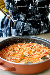 Unrecognizable person pouring olive oil in pot of orzo pasta, with tomato sauce, vegetables and seafood. Selective focus.