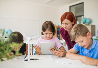 Group of small school kids with teacher sitting in circle in class, using tablet.