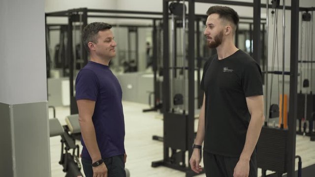 Young professional fitness trainer and middle-aged sportsman shaking hands standing in front of fitness equipment in the gym. Concept of sport, active lifestyle, healthcare
