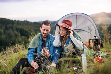 Young tourist couple travellers with tent shelter sitting in nature, using smartphone.