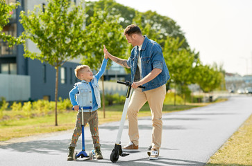 family, leisure and fatherhood concept - happy father spending time with little son riding scooters...