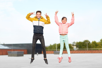 childhood, happiness and fun concept - happy children or brother and sister jumping on rooftop and showing peace gesture