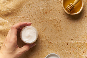 Spa beauty skincare flatlay on travertine background wiith female hand holding a  small container of cream. Overhead view, copy space