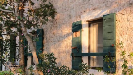 Close-up of a one-storey old stone house in the traditional Balkan national style with open wooden shutters. Coniferous trees surround the house