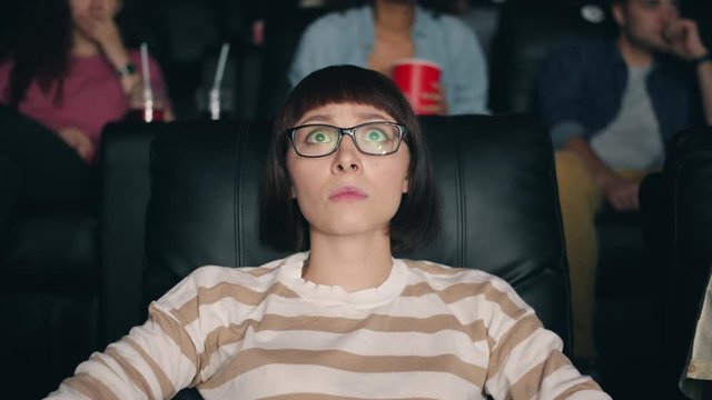 Frightened girl in glasses watching scary movie in cinema closing eyes with hands sitting in comfortable seat in dark room. People, emotions and youth concept.