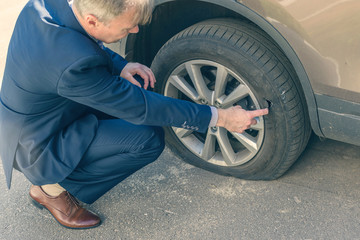 Obraz na płótnie Canvas A businessman in a blue suit crouching near his car and checks the degree of damage to a punctured wheel. Hole in the tire. Concept