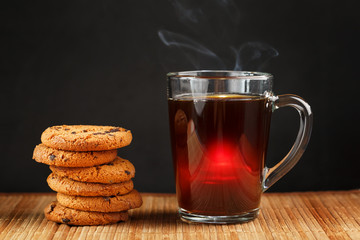 A pile of oatmeal cookies with chocolate chips and a mug of fragrant black hot tea in on a bamboo substrate, on a dark background. Handmade cookies for a healthy breakfast
