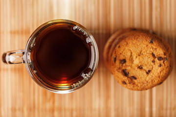 A pile of oatmeal cookies with chocolate chips and a mug of fragrant black hot tea in on a bamboo substrate, on a dark background. Handmade cookies for a healthy breakfast.