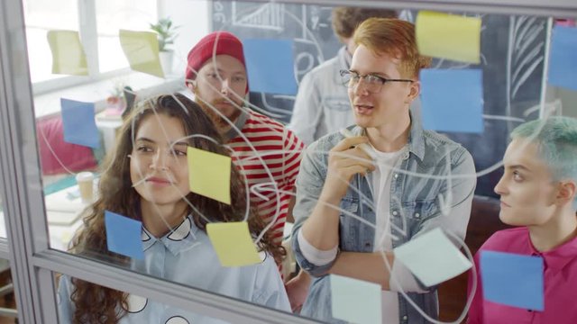 Waist-up shot of red-haired young Caucasian man standing in front of glass wall with sticky note diagram in office, colleagues coming over, discussing new idea together and nodding enthusiastically