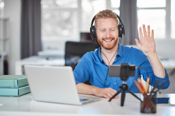 Hardworking employee having video call over smart phone while sitting in office and waving. On head are headphones and on desk laptop. Start up business concept.