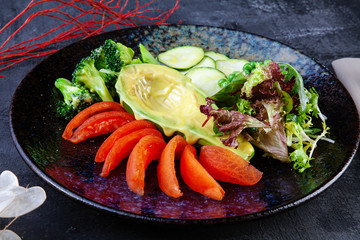 Close up view on green salad with avocado in served in black textured plate on dark background. Flat lay food. Vegan fresh salad. Copy space for design