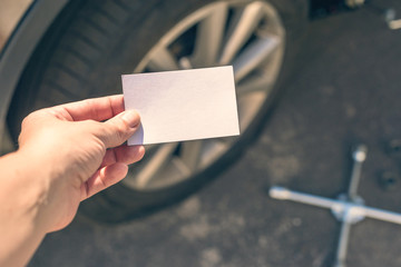 woman crouching near her car with  punctured wheel. girl holding business card with data of isurance company. Concept. mock up - obrazy, fototapety, plakaty