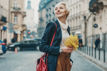Outdoor portrait of young happy smiling lady with braces on her teeth walking in street of European city, holding bouquet of autumn leaves. Copy, empty space for text