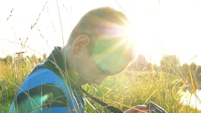 little boy takes pictures with a digital camera and opens the folding screen of the camera in the grass at sunset