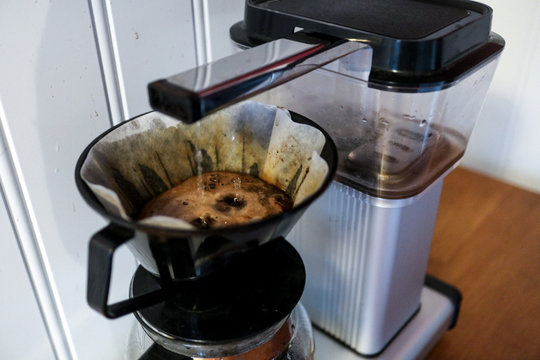 A coffee maker i standing on a wooden kitchen counter while the hot water is hitting the powder before the liquid hits the pot.