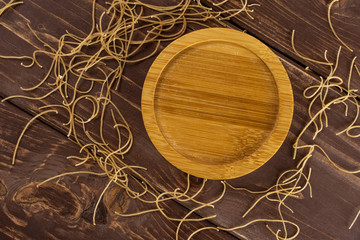 Lot of pieces of thin raw pasta noodles with empty bamboo plate flatlay on brown wood