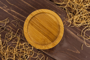 Lot of pieces of thin raw pasta noodles with empty bamboo plate in centre flatlay on brown wood