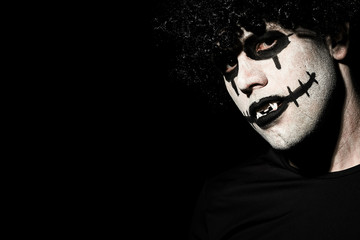 The terrible creepy clown in a black wig grins, looking at the camera. Empty space on the left, black background.