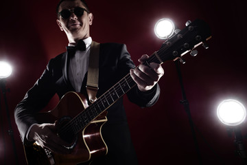 Fototapeta na wymiar Portrait of classical musician with guitar in red studio with stage lighting. Guitarist in black glasses and suit with a bow tie improvises on instrument