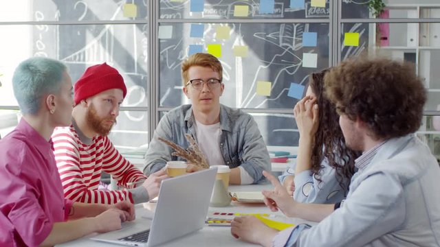 Medium shot of five young Caucasian creative men and women sitting in studio around table, brainstorming for new project, proposing ideas, discussing and arguing