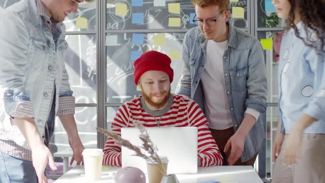 Chest-up shot of young red-haired Caucasian hipster in hat and stripy sweatshirt sitting in creative studio and working on laptop, colleagues joining, looking at screen together and discussing project