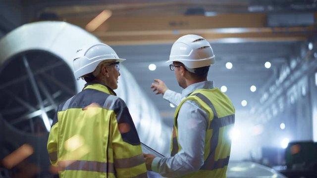 Two Heavy Industry Engineers Stand in Pipe Manufacturing Factory, Use Digital Tablet Computer, Have Discussion. Construction of Oil, Gas and Fuels Transport Pipeline. Back View Sparks Flying