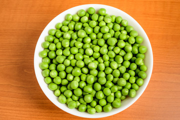 Freeh organic green peas in a white round bowl on a wooden table in soft focus, top view