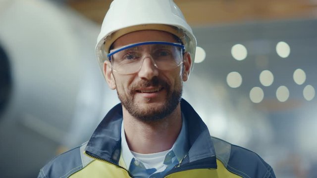 Portrait of Professional Heavy Industry Engineer / Worker Wearing Safety Uniform, Goggles and Hard Hat Smiling. In the Background Unfocused Large Industrial Factory where Welding Sparks Flying