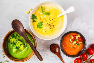 Three different vegetable cream soups in bowls on gray background. Corn, cucumber and gazpacho...