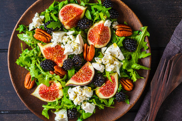 Salad with figs, feta cheese and blackberries in a wooden plate on dark background, top view.