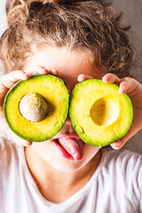 Healthy baby food concept. Girl child holds sliced avocado in place of the eyes.