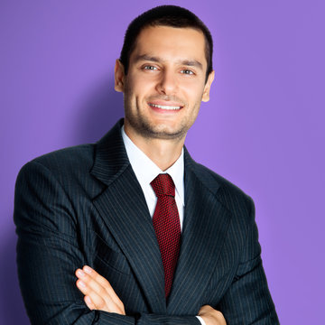 Portrait of happy confident business man in black suit and red tie, with crossed arms pose, standing against violet color background. Square composition picture.