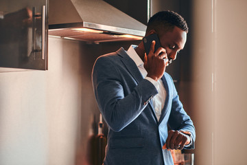 Young african man in white shirt is chatting by mobile phone at the kitchen.