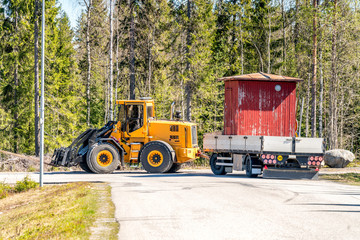 Big yellow tractor relocates old small red woden house in the trailer, sunny summer day, nearby to Umea city, North Sweden