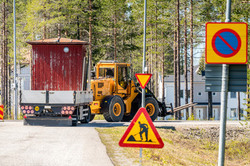 Big yellow tractor turn right while relocates old small red woden house in the trailer, sunny summer day, nearby to Umea city, North Sweden.Road construction sign at the road side