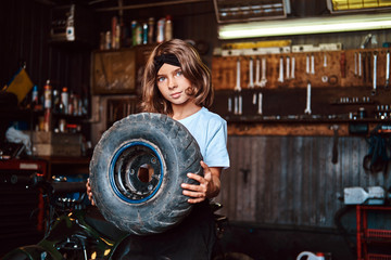 Fototapeta na wymiar Nice small girl is posing for photographer with big wheel from car at auto service.
