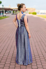 Smiling dreamy Caucasian woman in amethystine floor-length dress turned her back on pavement.