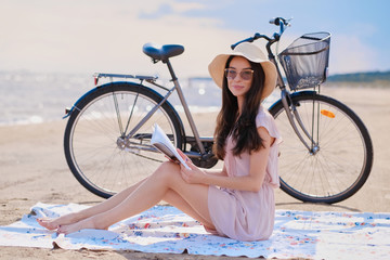 Beautiful cheerful brunette in hat is enjoying summertime while reading book at the sea next to her bike.