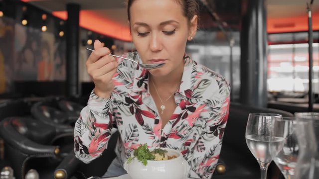 Beautiful female tourist in a colorful shirt spoon eats a traditional French onion soup with croutons and greens in a Paris restaurant. Slow Motion.