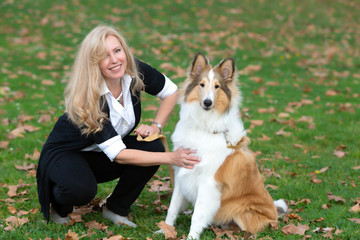 Blond Caucasian woman is sitting on a green grass and fall foliage in a park with collie dog