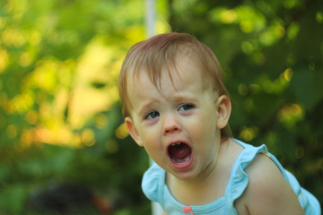 Fototapeta premium little screaming white blond-haired child on a background of blurry green foliage