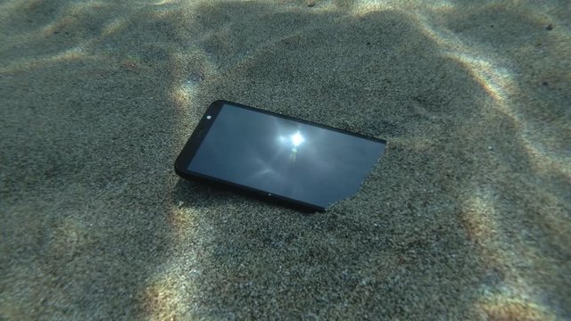 Smartphone lies on a caved seabed on its screen reflects the sun with rays and surface of blue water with small waves. Underwater view. Mediterranean Sea, Europe.